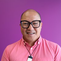 A man with a pink shirt is wearing glasses. He is in front of a purple background. 
