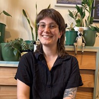 A person smiling. They are wearing glasses and have tropical pot plants behind them.