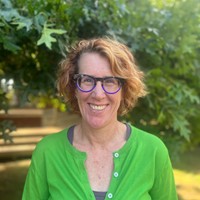 A woman smiling. She has a green cardigan and is surrounded by trees. 