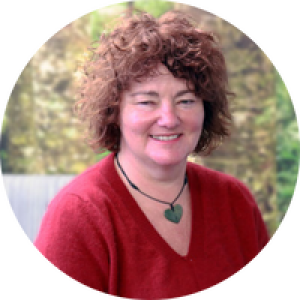 Amanda sits in front of a forest scene. She has curly brown hair, a bright red v neck top and a greenstone pendant the shape of a heart is around her neck