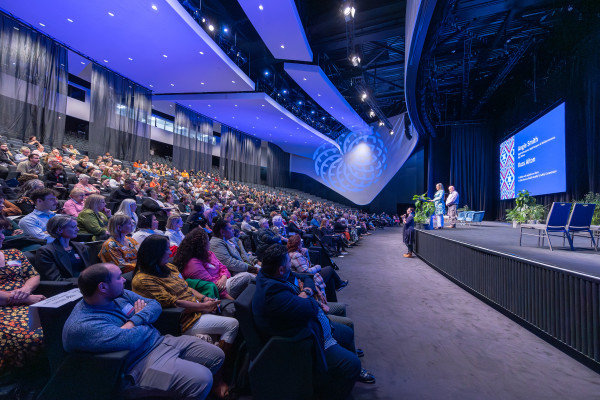 An audience of four hundred people are sitting in the auditorium at Te Pae Christchurch Convention Centre for the Our voices | Ō mātou reo forum. They are listening to Te kāhui mahi ngātahi co-chairs Angie Smith and Russ Aiton as they set the scene for the day. There is blue stage lighting above them and lush and greenery on stage next to the lectern.