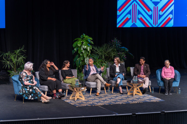Community panel ‘Out of the box’ facilitated by Arrun Soma. Sitting left to right: Joanne Neilson, Wikitoria Kurene, Lisa Lawrence, Arrun Soma, Jodie Bennett, Vishal Rishi and Jodie Bennett. They are sitting on comfy chairs in a row onstage and looking at each other.