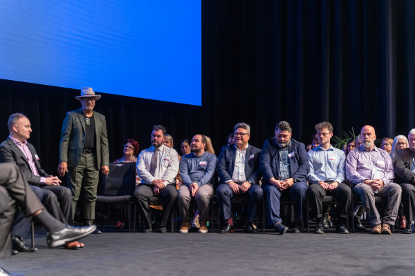 On stage Doug Edwards Te Pou Tūmatanui, Te Tāhū Hauora responds to the mana whenua Te Taumata Tapu o Ngāi Tūāhuriri with a mihi. Te Tāhū Hauora staff sitting next to him. Thirty Te Tāhū Hauora staff are seated on stage opposite mana whenua. Tane are sitting and the front and wāhine are at the back. They are listening to the response.