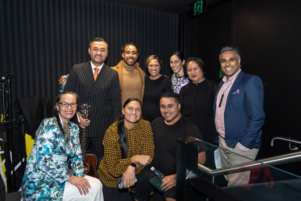 People are sitting backstage at the Our voices forum at Te Pae Christchurch Convention Centre. They and are smiling at the camera before they go on stage. The people in the photo are: Front row, left to right: Angie Smith, Dame Valerie Adams, Matthias MacKay. Back row, left to right: Francis Tipene, Moses MacKay, Kaiora Tipene, Lauagaia Jeffries, Edna Tu’itupou-Havea and Arrun Soma.
