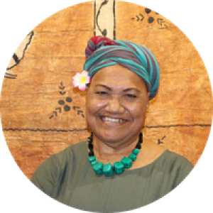 Person wearing a patterned headwrap, a flower accessory, and a teal beaded necklace, photographed in front of a textured tapa‑style background.