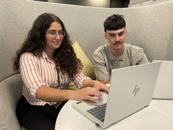 Two interns sit together in a modern office booth, working on laptops. The intern on the left has long, curly hair and is wearing a striped shirt and a pounamu pendant. The other intern has short hair and is wearing a patterned collared shirt. They are seated at a small round table with laptops open in front of them.