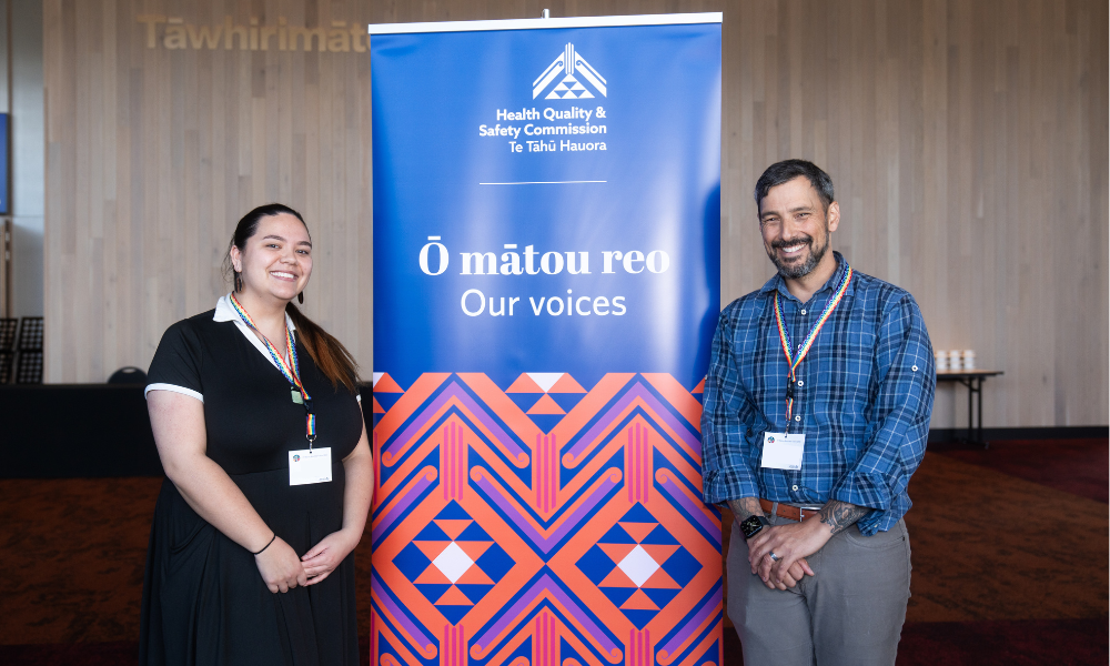A woman and man stand smiling beside a banner. The banner reads: Ō Mātou Reo Our Voices. It is navy blue, and has an orange and purple tohu design on the bottom. The woman stands on the left. She has long dark hair and wears a black top and skirt, with a rainbow lanyard. The man is on the right of the banner, he has short dark hair and a beard and wears a blue checkered shirt with grey pants. 