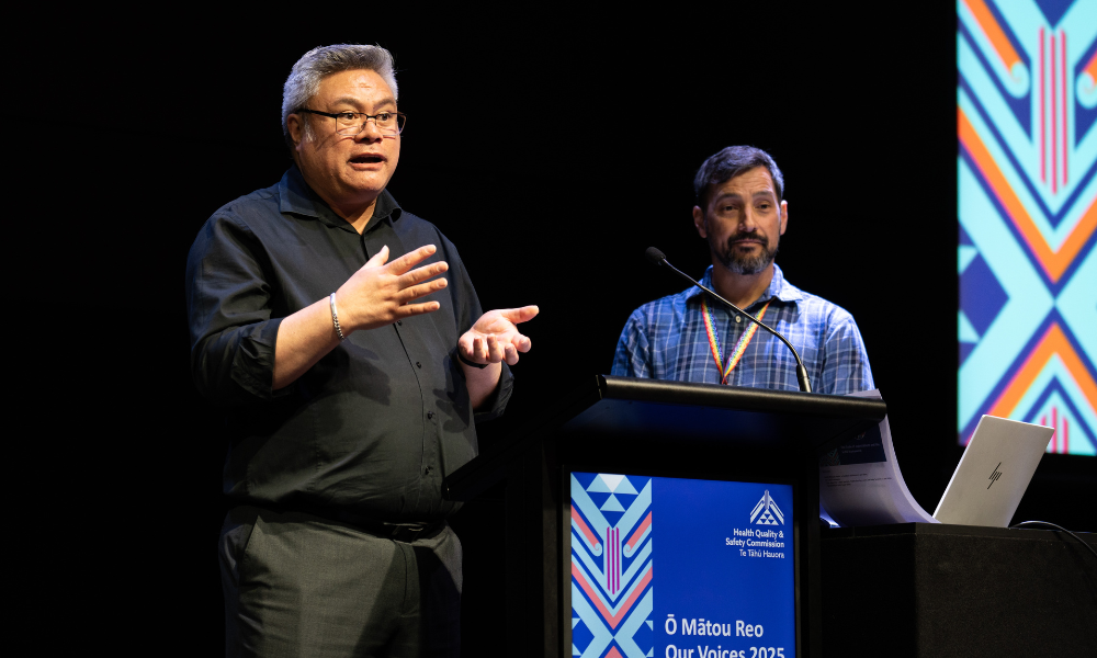 Two men are giving a presentation from behind a podium. The man on the left has short, grey hair. He wears glasses, a black shirt and grey pants. He is speaking and gesturing with his hands. The man on the right has short, dark hair. He wears a blue shirt and rainbow lanyard. He is listening, with a focussed look on his face.