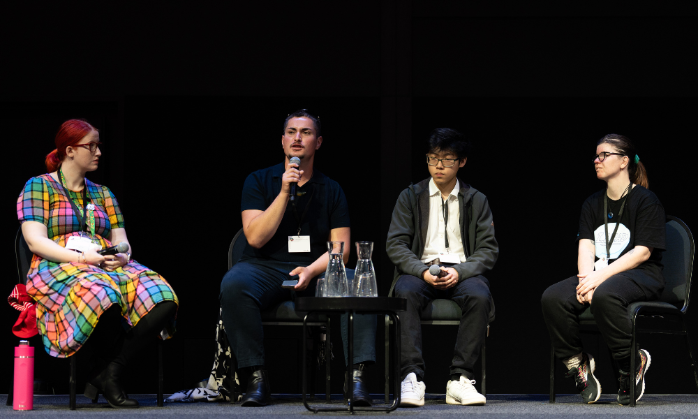 Four members of the Youth Voices panel sit on stage. From left to right, there is a woman wearing a colourful, checkered dress with short red hair, the man to the right is speaking into the microphone, he wears dark clothing and has short hair. The next to the right is a young man who is sitting listening. He wears a light shirt, dark cardigan and glasses. Next to him is a young woman with long hair tied in a pony tail. She wears a dark top and black jeans.