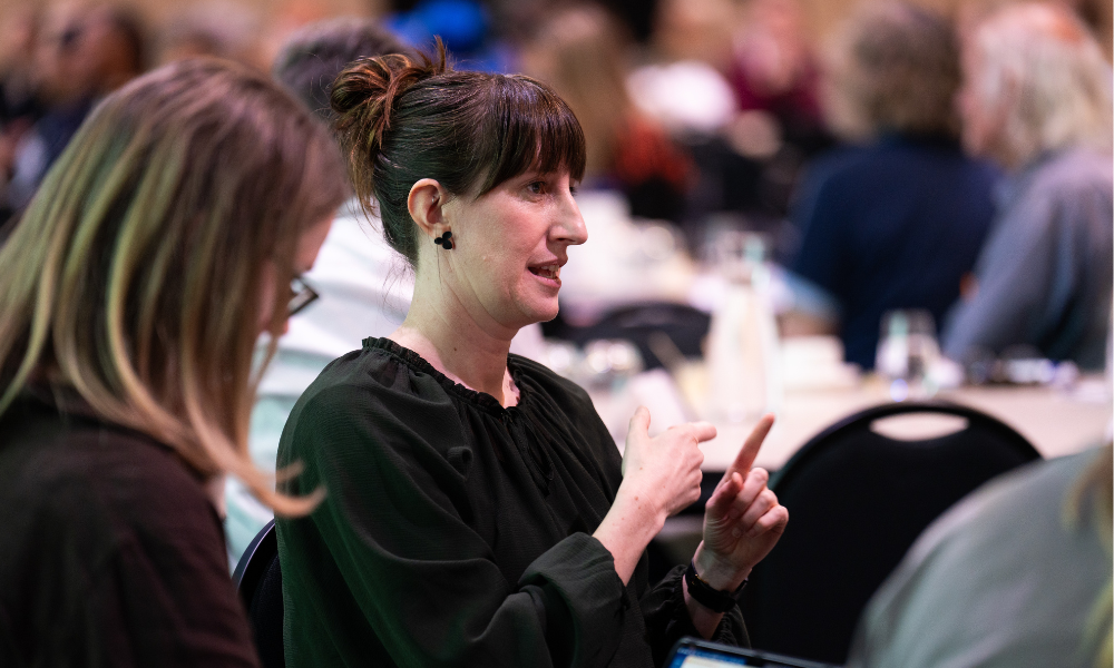 A photo of a sign language interpreter translating for deaf participants at a table. She has dark hair pulled up into a bun, and a horizontal fringe. She wears a black top.