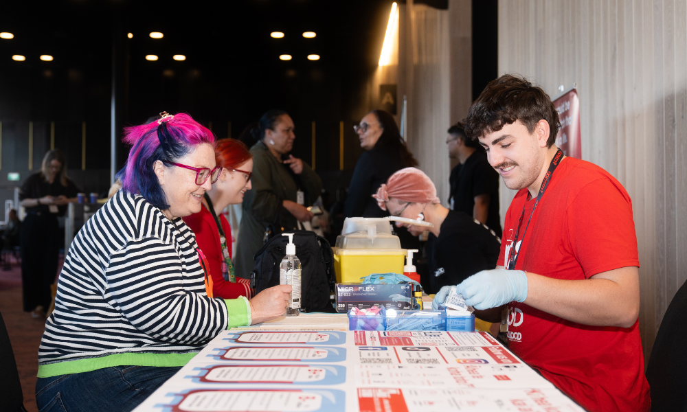 Two people sit at a promotional table, looking at information in front of them and smiling. A woman sits on the left of the table. She has pink and black hair, a stripy top and blue jeans on. She also wears red glasses. A man sits across from her with short, dark hair, a dark moustache, red t-shirt and light blue gloves on.