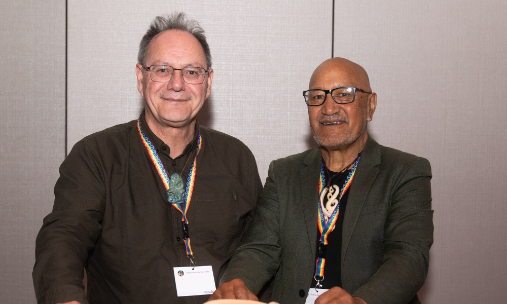 Two men pose for the camera.  The man on the left has short hair and wears glasses, a dark green shirt, rainbow lanyard and hei tiki around his neck. The man on the right has no hair, he wears glasses, a black shirt, green blazer with rainbow lanyard and carving around his neck. Both men are smiling at the camera.