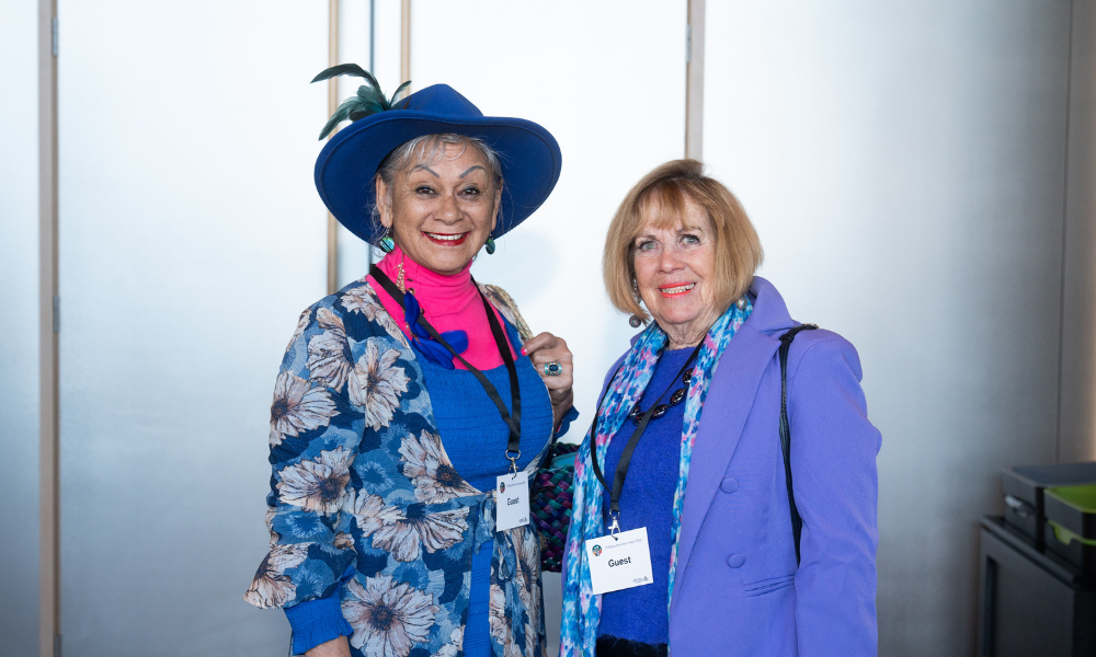 Two women pose for a photo. They are looking straight at the camera and smiling. The woman on the left wears a blue rimmed hat with a feather in the top. She has a bright pink top on under a blue dress, with a patterned blue cardigan. The woman on the right has short blonde hair and a blue top with a purple jacket and multicoloured scarf.