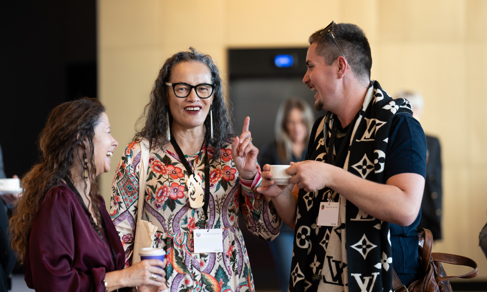 Three people are talking and laughing. The woman on the left has long hair and wears a purple top. The woman in the middle wears a colourful dress, with long hair and glasses. She has her finger raised in the air and is smiling. The main on the right is holding a cup of tea or coffee. He is wearing a scarf and has sunglasses on his head.