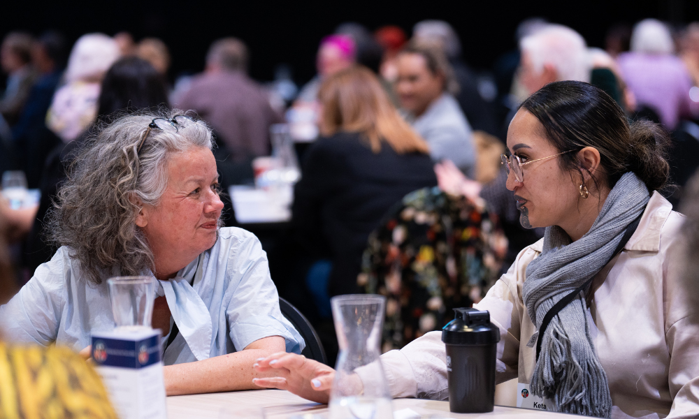 Two women are sitting at a table, talking. The woman on the left has grey hair with glasses on her head. She is wearing a blue shirt. The woman on the right has dark hair, glasses, a moko kaue and she is wearing a grey scarf and beige top. They are deep in conversation, with focussed looks on their faces. They are sitting in a busy room, behind them are many other people sitting and talking. 