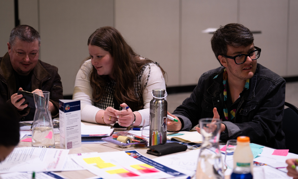 Three people are sitting at a table during a group activity. In front of them, the table is covered in post it notes, notepads, pens and waterbottles. The person on the left is looking at his phone. He has short hair and a dark top on. The woman in the middle has long hair and a white top on. She is also looking at the man’s phone.  The third man on the right is wearing glasses and a dark top. He is looking in the other direction, talking to someone out of frame. 