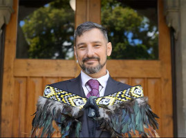 Carlton has silvery grey short hair and a beard. He is wearing a navy suit, purple shirt and purple tie. He has a pounamu around his neck and is adorned with a kākahu