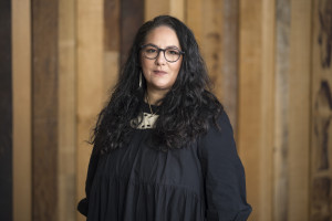 Cherie Seamark, a wāhine Māori, has long black curly hair and glasses. She is wearing a black blouse and has a bone carving hanging around her neck