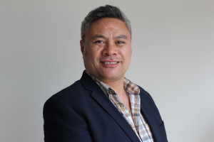 DJ, a Māori man, has closed cropped silver and grey hair. He is wearing a check shirt and a navy blue suit jacket