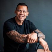 Dave, a Māori and Samoan man, has short cropped black hair and native design sleeve tattoos on both arms. Dave is wearing a black t-shirt and sitting backwards in a chair