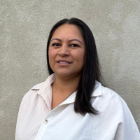 Hera, a wāhine Māori, has long straight black hair and is wearing a white button up collared short