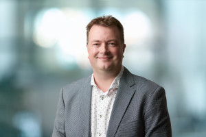 Justin has short blond hair. He is wearing a white shirt with black spots and a grey pinstriped suit jacket