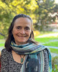 Kaaren has straight should length brown hair. She is wearing a black and white patterned blouse, a pounamu and a multi coulored scarf