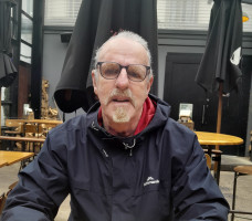 Martin has short white hair, moustache and beard. He is sitting at a table in a cafe, wearing a dark blue navy rain jacket 
