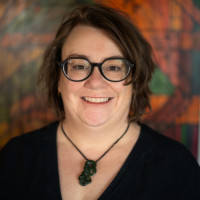 Moira a wāhine Māori has brown tousled hair down to the bottom of her neck. She is wearing a black top and black rimmed glasses and has a carved pounamu around her neck