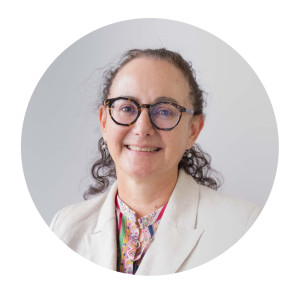 Nicole has long curly brown hair tied back from her face, and tortoiseshell glasses. She is wearing a cream blazer over a multi coloured blouse