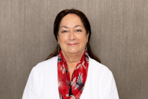 Norma has long dark hair behind her shoulders. She is wearing a white top with a red scarf patterned with piwakawaka fantail