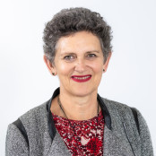 Rae has short wavy grey hair. She is wearing a grey jacket over a red patterned blouse, a black necklace and earrings, and lipstick that matches her blouse