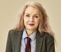 Rose has wavy long blonde hair and piercing blue eyes. She is wearing a blue shirt and a maroon tie with a grey marle blazer