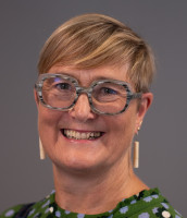 Sarah is smiling at the camera. She wears grey/white eyeglasses, white earrings and a green top.