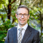 Sir Ashley has short greying hair and glasses. He is wearing a white shirt, grey check tie and blue check suit jacket. He stands in front of tree foliage