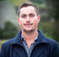 Stanley has close cropped brown hair and facial hair. He is wearing a blue patterned short with a navy blue coat over the top