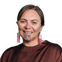 Tanya, a wāhine Māori, has brown hair pulled back off her face and a moko kauae. She is wearing a brown top and has long dangling earrings with intricate fern patterns