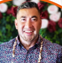 Zecheriah has short grey hair and facial hair. He is wearing a colourful floral patterned shirt and has a traditional shell necklace