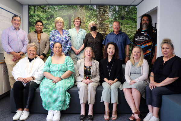 An image of a group of people. One row in the front is sitting and the other row stands behind them. They are smiling at the camera and standing in front of a forest background.