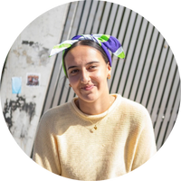 Kya is sitting on a bench in front of a grey wall. She is wearing a yellow knitted sweater and has a heart shaped pendant necklace. Kya has dark hair tied back with a purple, green and white scarf. 