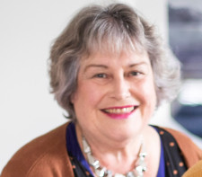 Bernadette has short grey hair and bright red lipstick. She is wearing a brown cardigan over a patterned top and a beaded necklace