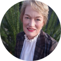 A head and shoulders photo of Clare Perry. Clare has short blonde hair.  Clare is wearing red lipstick, a white blouse with a frilly neckline and a checkered blazer