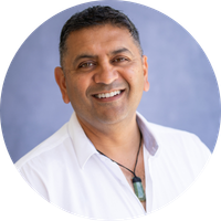 Rakesh has short dark hair and is wearing an open neck collared white shirt and a pounamu