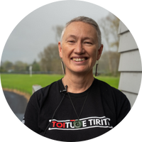 Taima has very short grey hair and is wearing dangling greenstone earrings. She has a black t-shirt with Toitū te tiriti printed on the front and a greenstone around her neck