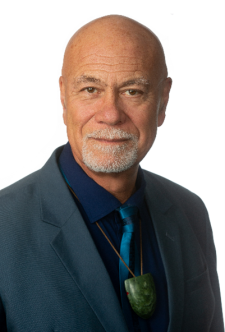 Denis is wearing a dark suit jacket, blue shirt and tie and a greenstone around his neck. Denis has a grey beard and moustache 
