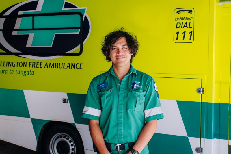 A man in a green paramedic uniform stands in front of a bright yellow Wellington Free Amulance