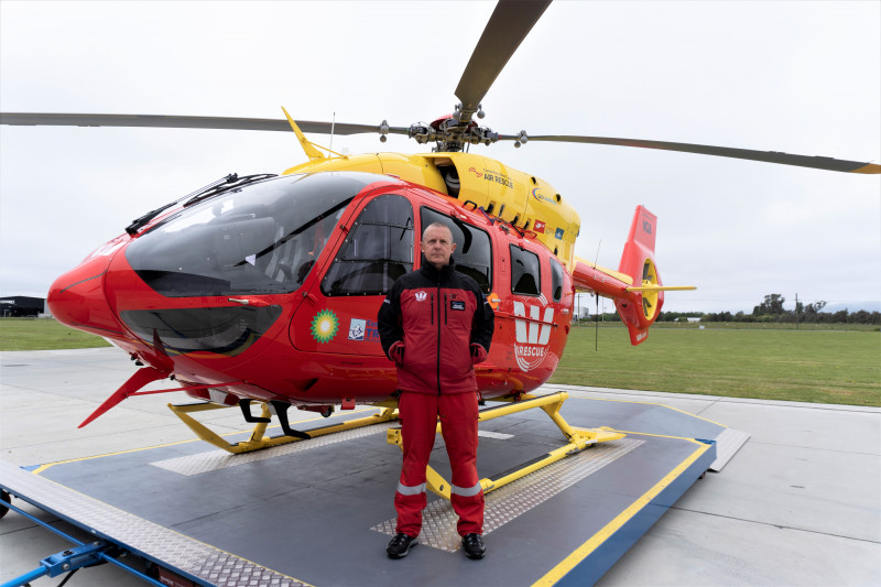 A paramedic dressed in a red uniform. He is standing in front of a red and yellow helicopter with Wespac written on it. 