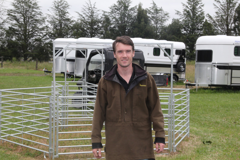 Thomas stands in front of a stock pen and two caravans. He's wearing a brown hunting and fishing jacket.