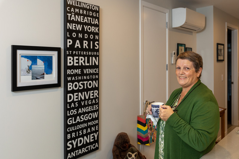 A woman in her 60s (Cheryl) standing in her home next to a large artwork with a series of countries written on it in large white letters, from Wellington to Antarctica. She is holding a cup of tea and smiling. 
