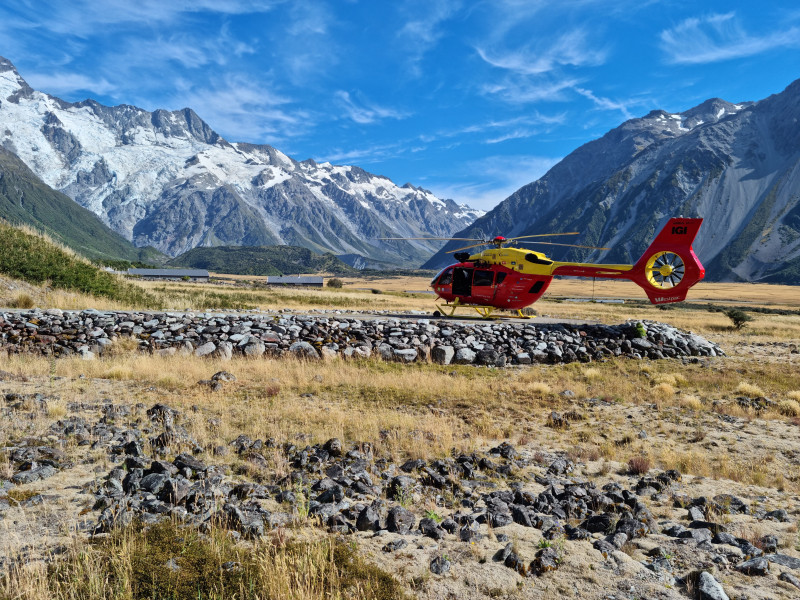 A red and yellow helicopter sitting in a rocky field with snow capped mountains in the background. 